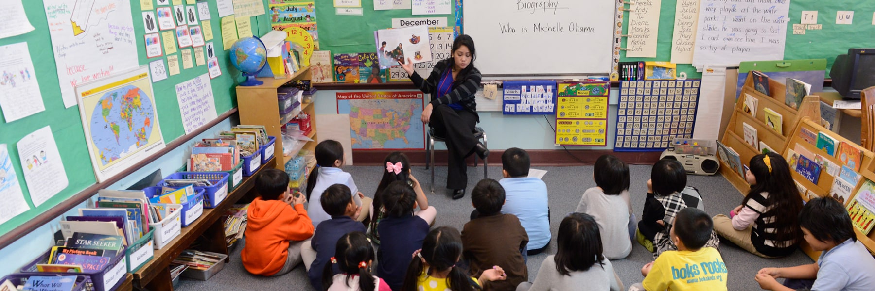 young teacher holding up a book and seated in front of large classroom of small children