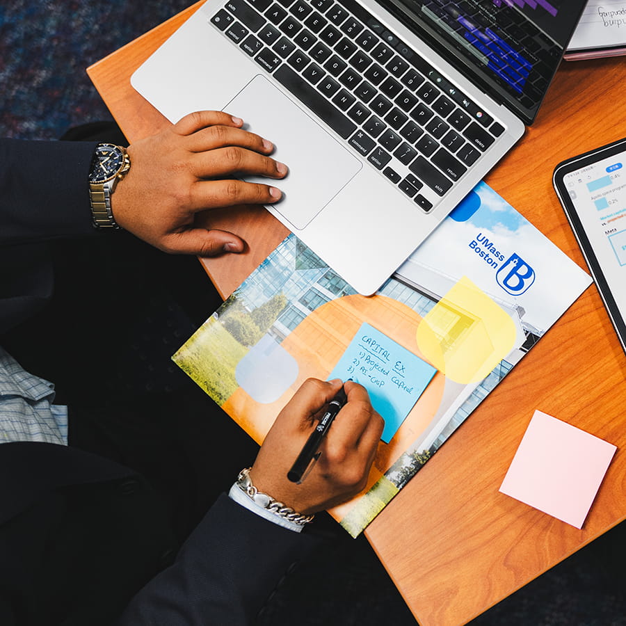 close up hand wearing wristwatch on laptop making notes on sticky pad over the UMass Boston brochure
