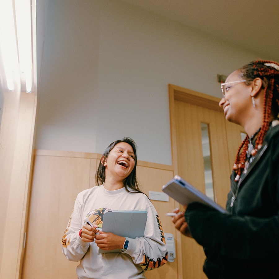 Two students laugh in classroom holding laptops near a whiteboard.