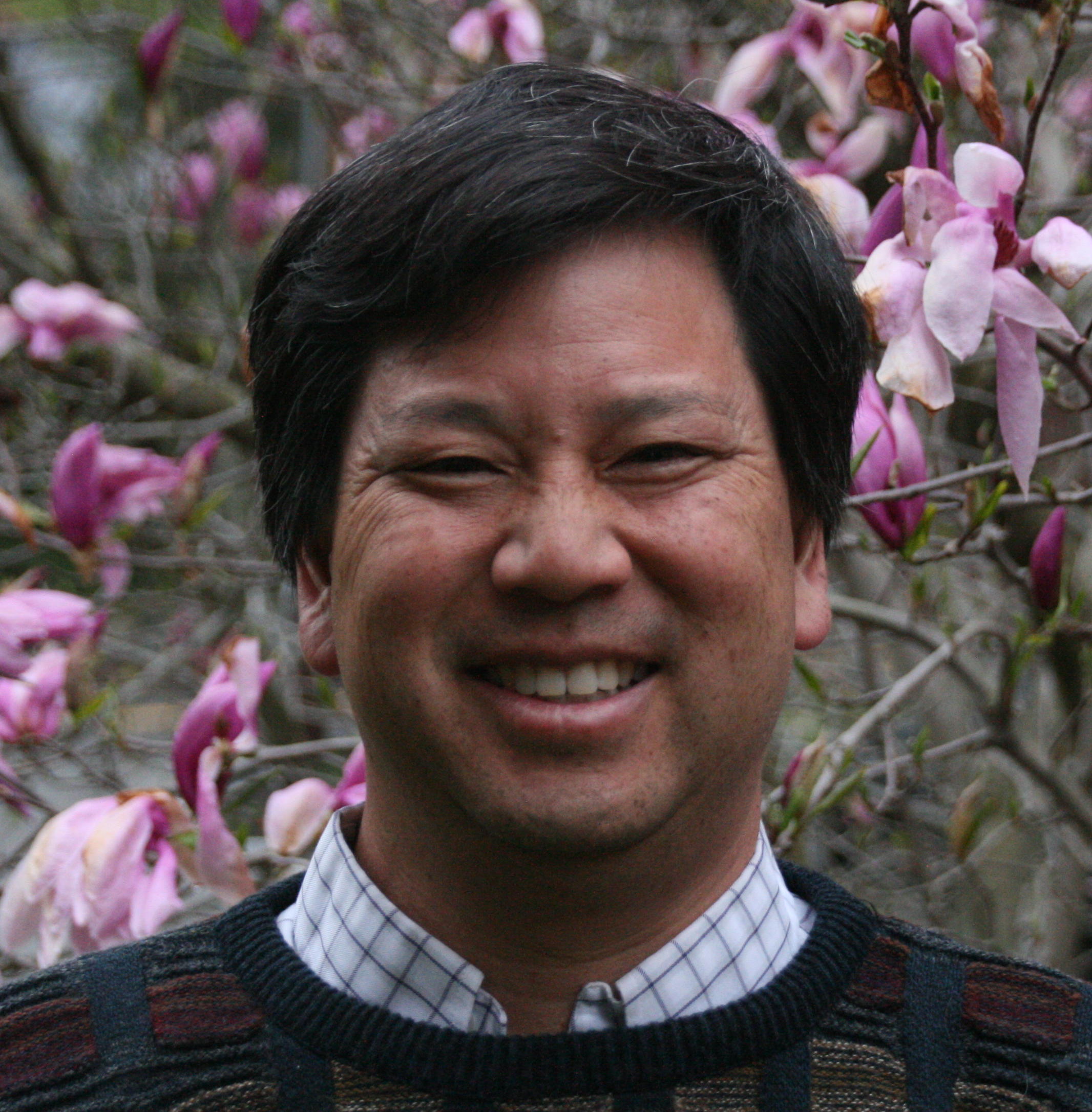 man with short dark hair and dark sweater in front of pink flowers