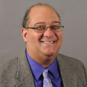 Man with hair pulled back, metal frame glasses, purple shirt and gray blazer against gray background