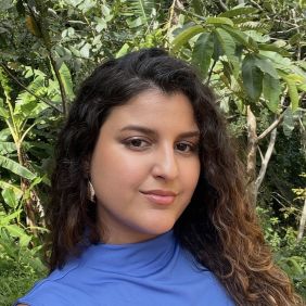 woman with long brown curly hair and blue mock turtleneck in front of tropical plants