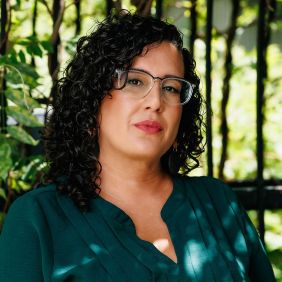 Woman with curly dark hair and glasses wearing emerald green, leaves behind her