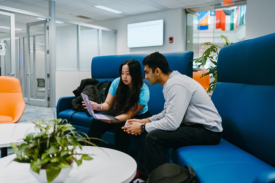 two students sit in couch in One Stop office looking at screen on laptop